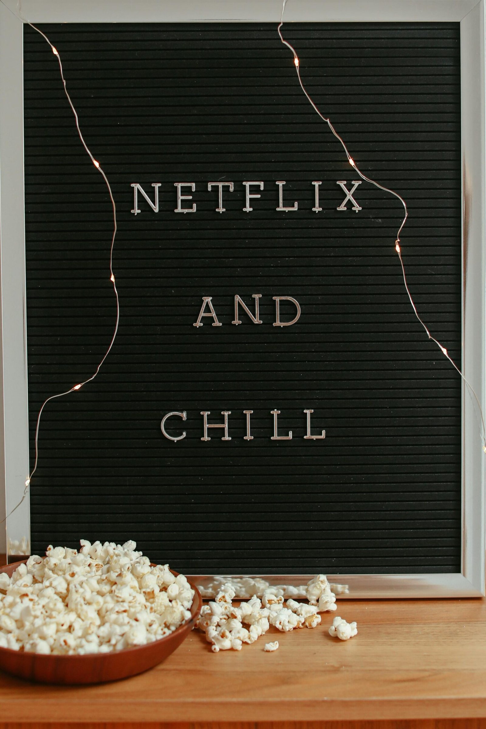 Letter board with 'Netflix and Chill' text, surrounded by popcorn and string lights on a wooden surface.